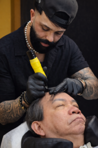 Scalp micropigmentation (SMP) specialist performing hairline restoration treatment on a male client using a professional SMP machine in a clinic setting.